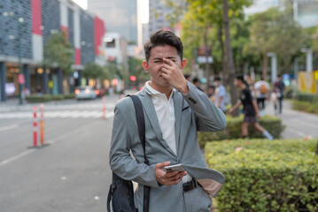 A young asian man suffers from sinusitis or runny nose while walking around the city. Urban air...