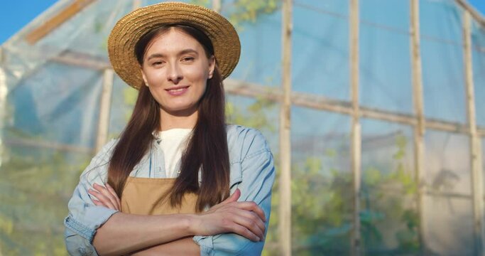 Portrait of young farmer in straw hat. Attractive young woman poses with arms crossed against background of greenhouse. Female farmer outside smiling and looking at camera.