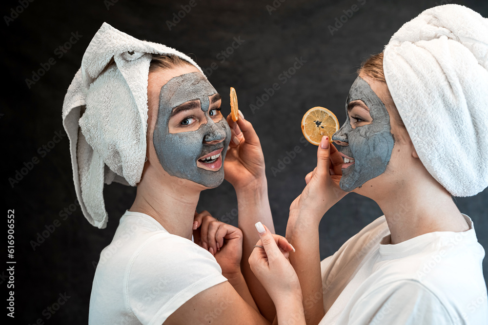 Wall mural two young women in white towel made beauty clay facial mask and hold orange isolated on black.