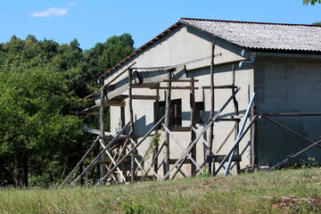 Industrial storage hangar with two small windows and homemade wooden scaffolding prepared for facade reconstruction on side of small hill covered with uncut grass next to dense forest on warm sunny