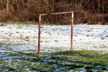 Rusted old goal post without net left on abandoned children playground covered with freshly fallen snow mixed with uncut grass filled with multiple molehills next to dense forest vegetation