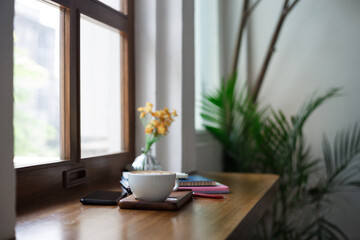 Close-up view, white coffee cup with latte coffee put on wooden counter and smart phone, note book put on digital tablet by window, coffee break and relax after work in cafe
