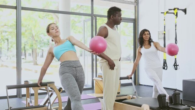 Hispanic woman exercising with pilates reformer and small fitness ball. African-american man pilates instructor assisting her with workout.