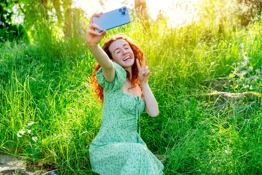 Outdoor portrait of happy red hair woman in green dress taking photos on mobile phone in the public park