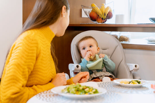 Mother looking at baby boy eating solid food on high chair at home