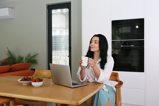 Thoughtful woman holding coffee cup sitting with laptop at table at home