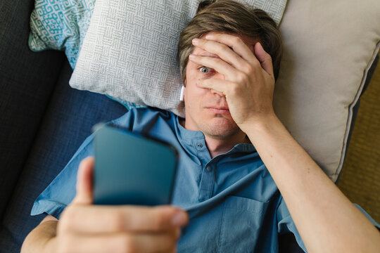 Worried man looking at smart phone lying on bed
