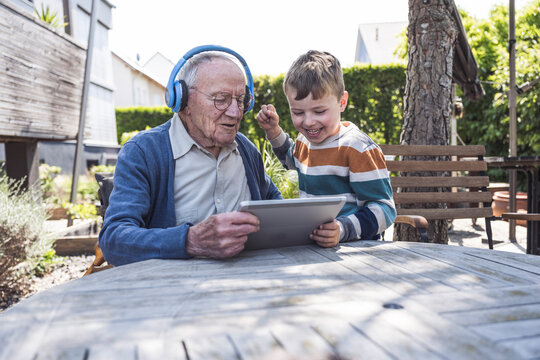 Senior man wearing wireless headphones using tablet PC with grandson