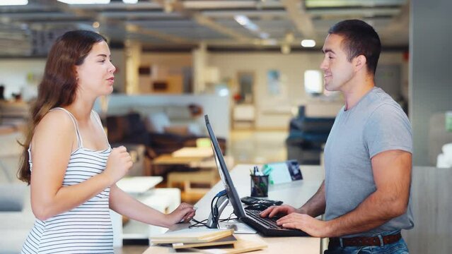 Focused young salesman of furniture showroom working with computer at information desk, advising female buyer on assortment . High quality 4k footage