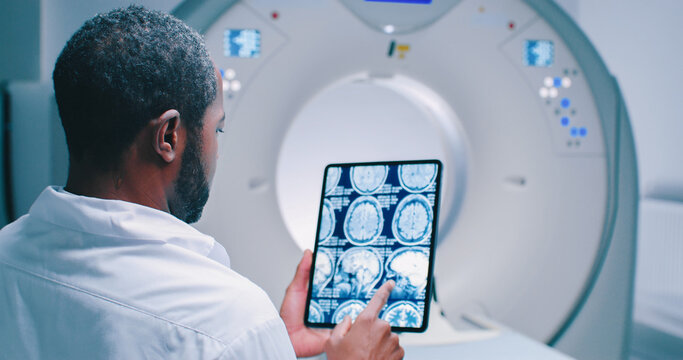 African American doctor examine scan attentively. Medical worker shooted from back. Doctor looks at MRI scan. Multicultural doctor is scrolling screen with magnetic resonance imaging scans of brain.