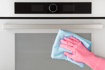 Woman hand in pink rubber protective glove using blue dry rag and wiping glass surface of oven door...