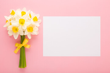 Bouquet of fresh white narcissus flowers with yellow ribbon and blank greeting card on light pink...