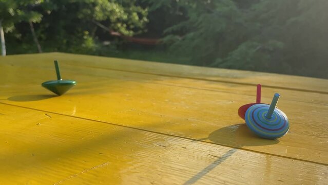 Close-up single spinning top green color spins on yellow table while other top toys fell outdoor. Rotating object as analogy for balancing life. Beautiful landscape park golden sun rays background