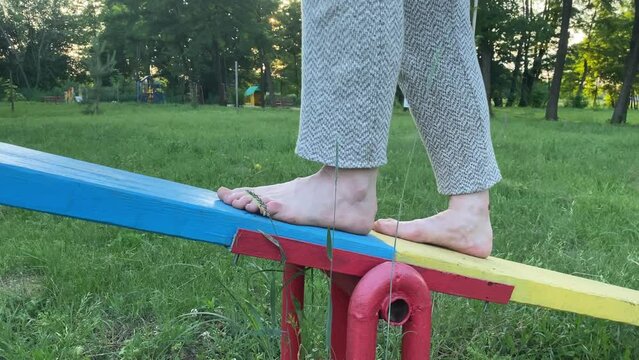 Barefoot woman walks on seesaw outdoors as analogy to balancing life, ascent and descent life cycle. Balance concept. Life work balance. Green landscape park in rays of sunset on background
