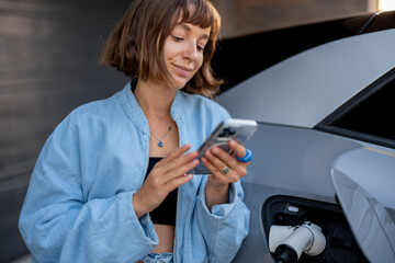 Young woman using smartphone leaning to her car and waiting while it charges. Concept of new technologies and modern lifestyle