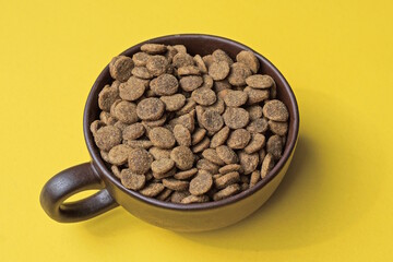 one ceramic cup full of round small brown cat food stands on a yellow table