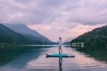sports woman stands on sup board. Paddle board on a beautiful lake at sunset. Sports on the water. Silence, carelessness and outdoor recreation. Beautiful sunrise landscape. Adventure in Italy Alps.  - obrazy, fototapety, plakaty