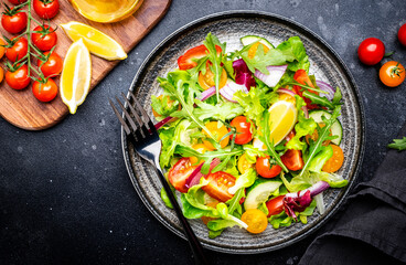 Summer vegan vegetable salad with colorful tomatoes, fresh cucumber, red onion, lettuce and arugula. Black table background, top view