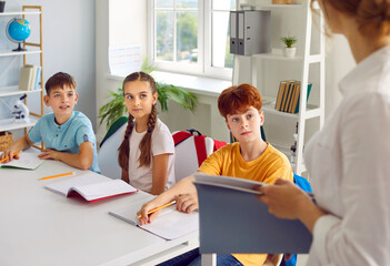 Cute smiling school-age children listen attentively to their teacher in spacious cozy classroom,...