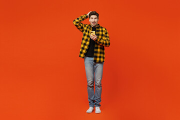 Full body scared sad uhappy young caucasian man he wear yellow checkered shirt black t-shirt hold head use mobile cell phone isolated on plain red orange background studio portrait. Lifestyle concept.