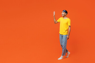Fototapeta premium Full body side view smiling happy young man wear pyjamas jam sleep eye mask rest relax at home walking go waving hand isolated on plain orange background studio portrait. Good mood night nap concept.