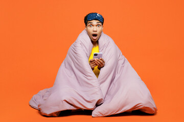 Full body amazed young man in pyjamas jam sleep eye mask sit wrapped in duvet rest relax at home use mobile cell phone isolated on plain orange background studio portrait Good mood night nap concept