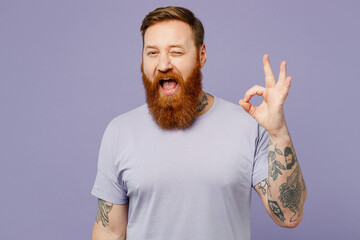 Young cheerful fun redhead bearded man wearing violet t-shirt casual clothes showing okay ok gesture wink blink eye isolated on plain pastel light purple background studio portrait. Lifestyle concept. - obrazy, fototapety, plakaty