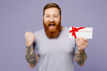 Young redhead bearded man wear violet t-shirt casual clothes hold gift certificate coupon voucher card for store do winner gesture isolated on plain pastel light purple background Lifestyle concept. - obrazy, fototapety, plakaty