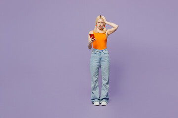 Full body scared sad young blonde woman she wear orange tank shirt casual clothes hold head use mobile cell phone isolated on plain pastel light purple background studio portrait. Lifestyle concept.