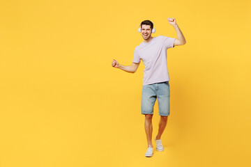 Full body young smiling cheerful man he wear light purple t-shirt casual clothes headphones listen to music dance raise up hands isolated on plain yellow background studio portrait. Lifestyle concept.