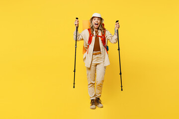 Full body shocked young woman carry bag with stuff mat hold trekking poles isolated on plain yellow background. Tourist leads active lifestyle walk on spare time. Hiking trek rest travel trip concept.