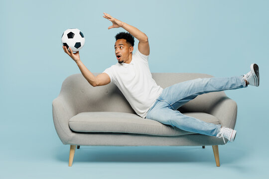Full body happy fun cheerful cool young man fan wear t-shirt cheer up support football sport team hold catch soccer ball lay down on grey sofa watch tv live stream isolated on plain blue background.