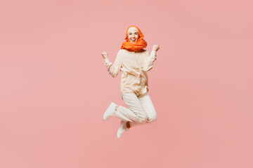 Full body young arabian asian muslim woman wearing orange abaya hijab jump high do winner gesture celebrate clenching fists say yes isolated on plain pink background. Uae middle eastern islam concept.
