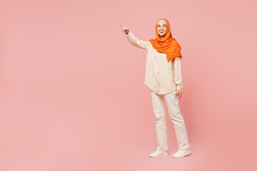 Full body smiling happy fun young arabian asian muslim woman wearing orange abaya hijab point index finger aside on area isolated on plain pink background. Uae middle eastern islam religious concept.