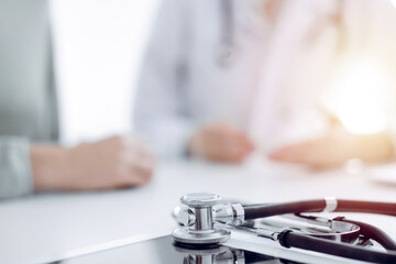 Doctor and patient are sitting and discussing something at the desk in the clinic office. The focus is on the stethoscope lying on the table, close up. Perfect medical service and medicine concept