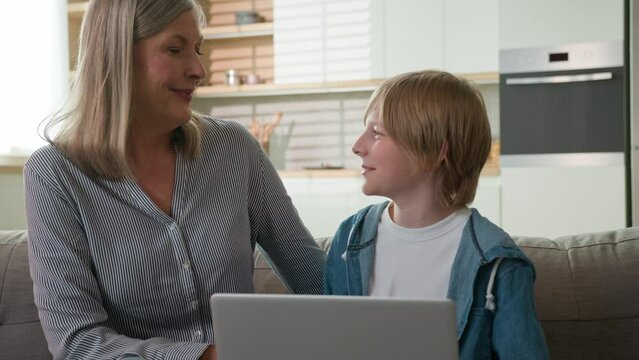 Happy smiling caucasian elderly woman grandmother and grandson grandchild boy use laptop rest on sofa at home together talking laughing embrace loving family hugging expressing tenderness relationship