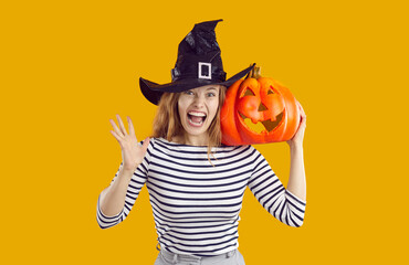 Funny woman on Halloween night. Studio portrait of happy beautiful young woman wearing black witch hat holding orange carved Jack o lantern pumpkin on her shoulder, looking at camera and screaming