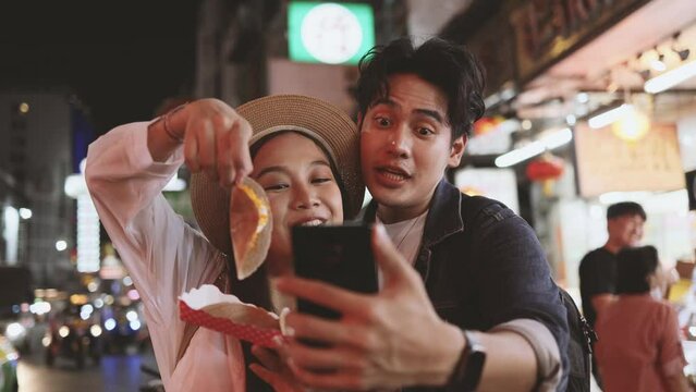 Asian couple tourist backpacker doing selfie and recording a video blog while eating food from street stall in night market with crowd of people at Yaowarat road, Bangkok