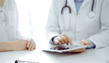 Fototapeta premium Doctor and patient sitting near each other at the desk in clinic. The focus is on female physician's hands pointing into tablet computer touchpad, close up. Medicine concept