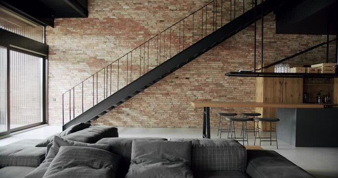Modern and spacious wooden kitchen room with wall bricks and black staircase and gray sofa in living room. Real Estate. Luxury kitchen and living room with wooden island and chairs.