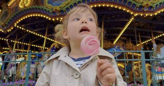 A three-year-old girl licks a colorful candy in an amusement park. Little girl eats pink candy at a festive children's event against the backdrop of lights. Children's holiday joy for children