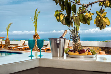 Relax and holiday concept, white bar counter with an ice basket containing a bottle, two blue glasses, and a fruit plate with bananas, apples, and pineapple.