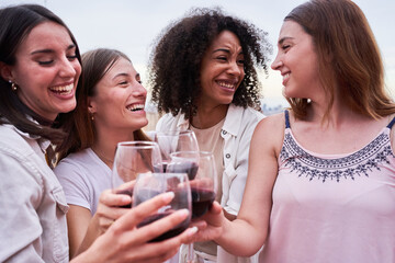 Four interracial female friends having fun together, toasting with red wine outdoors. Women enjoying on a rooftop celebrating and social gathering.