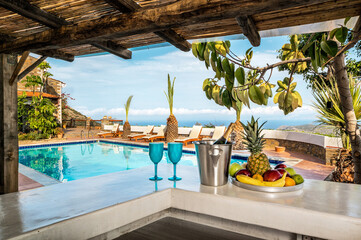 Relax and holiday concept, white bar counter with an ice basket containing a bottle, two blue glasses, and a fruit plate with bananas, apples, and pineapple. With the view to the pool. 