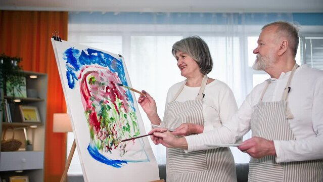 occupation in retirement, cheerful elderly spouses in aprons draw modern picture on canvas using brushes and paints at home