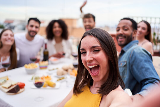 Group of multiethnic friends taking a selfie during social gathering in a rooftop. Cheerful photo of people dining outdoors in a terrace, looking at camera smiling.