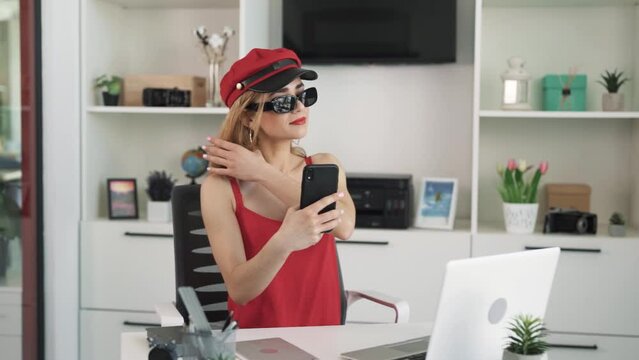 The young woman in a red dress and a red hat, wearing black glasses, takes a selfie on her phone, admiring herself on the camera.