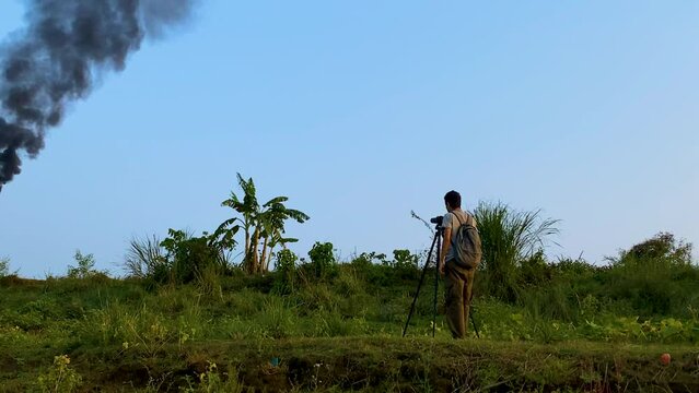 A documentary filmmaker, photographer is working on a pollution site and shooting a Petroleum refinery gas flare blazing fire pouring thick black smoke