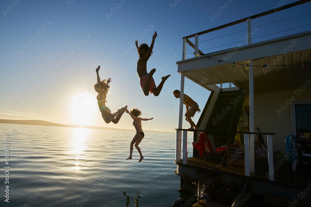 Wall mural young adult friends jumping off summer houseboat into sunset ocean - Wall murals