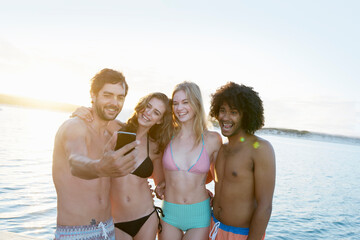 Young adult friends in bikinis swim trunks taking selfie at summer sunset ocean